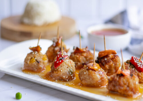 Vegan crispy fried rice balls served with sweet and sour sauce, garnished with sesame seeds and roasted red bell pepper, on a white plate with toothpicks for serving