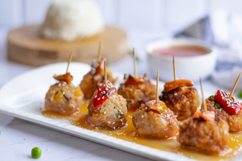 Vegan crispy fried rice balls served with sweet and sour sauce, garnished with sesame seeds and roasted red bell pepper, on a white plate with toothpicks for serving