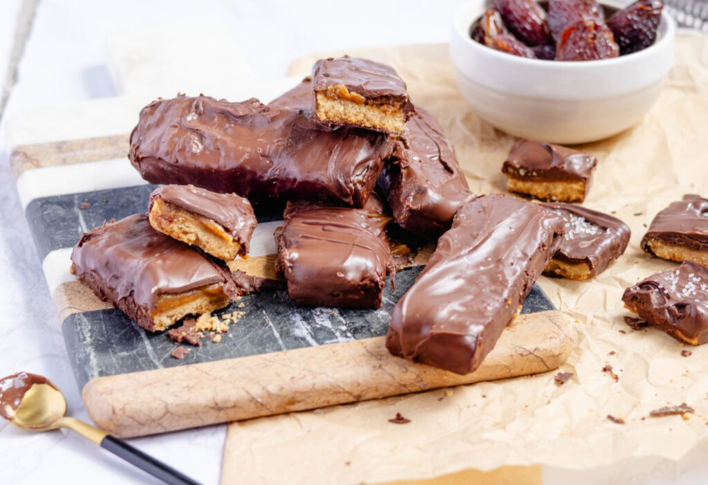 Close-up of homemade plant-based Twix bars in a candy mold, showing layers of almond flour shortbread, date caramel, and melted vegan chocolate on top.