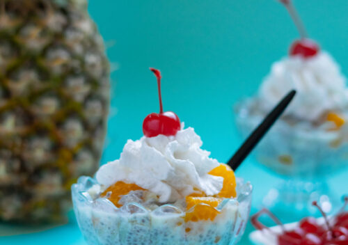 A creamy bowl of vegan and gluten-free frog eye salad made with chia seeds, mandarin oranges, and pineapple, styled over a blue backdrop with a pineapple in the background.