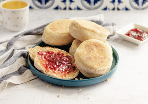 A plate of homemade vegan sourdough English muffins, one sliced open and spread with red jam, surrounded by a striped kitchen towel, with a mug of tea and extra jam in the background.