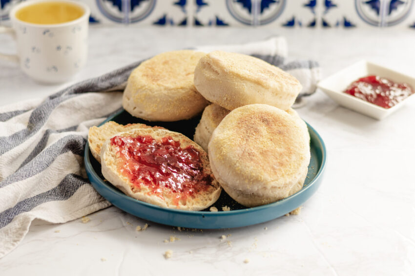 A plate of homemade vegan sourdough English muffins, one sliced open and spread with red jam, surrounded by a striped kitchen towel, with a mug of tea and extra jam in the background.