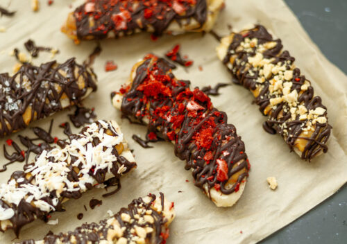 Close-up of frozen banana bars with peanut butter, dark chocolate drizzle, and a variety of toppings like crushed peanuts and freeze-dried fruit.