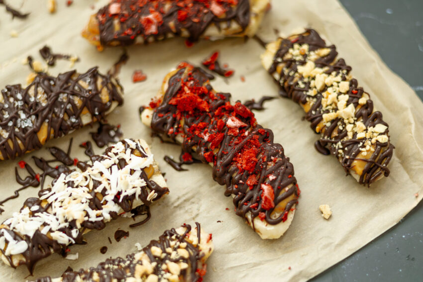 Close-up of frozen banana bars with peanut butter, dark chocolate drizzle, and a variety of toppings like crushed peanuts and freeze-dried fruit.