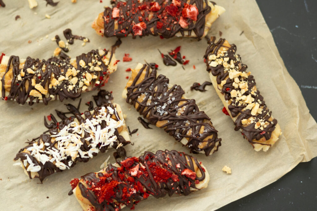 Frozen banana slices topped with peanut butter, drizzled with dark chocolate, and sprinkled with chopped peanuts, coconut, sea salt, and freeze-dried raspberries.