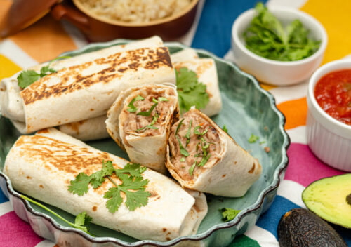 Crispy, golden-brown plant-based bean and cheese freezer burritos stacked on a green ceramic plate, garnished with fresh cilantro. Surrounded by salsa, avocado, and cilantro on a colorful tablecloth.