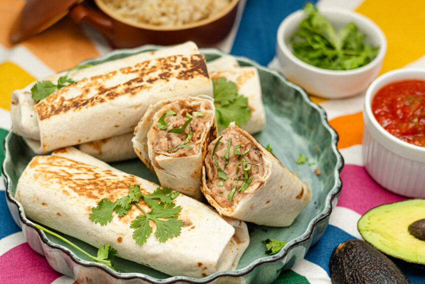Crispy, golden-brown plant-based bean and cheese freezer burritos stacked on a green ceramic plate, garnished with fresh cilantro. Surrounded by salsa, avocado, and cilantro on a colorful tablecloth.